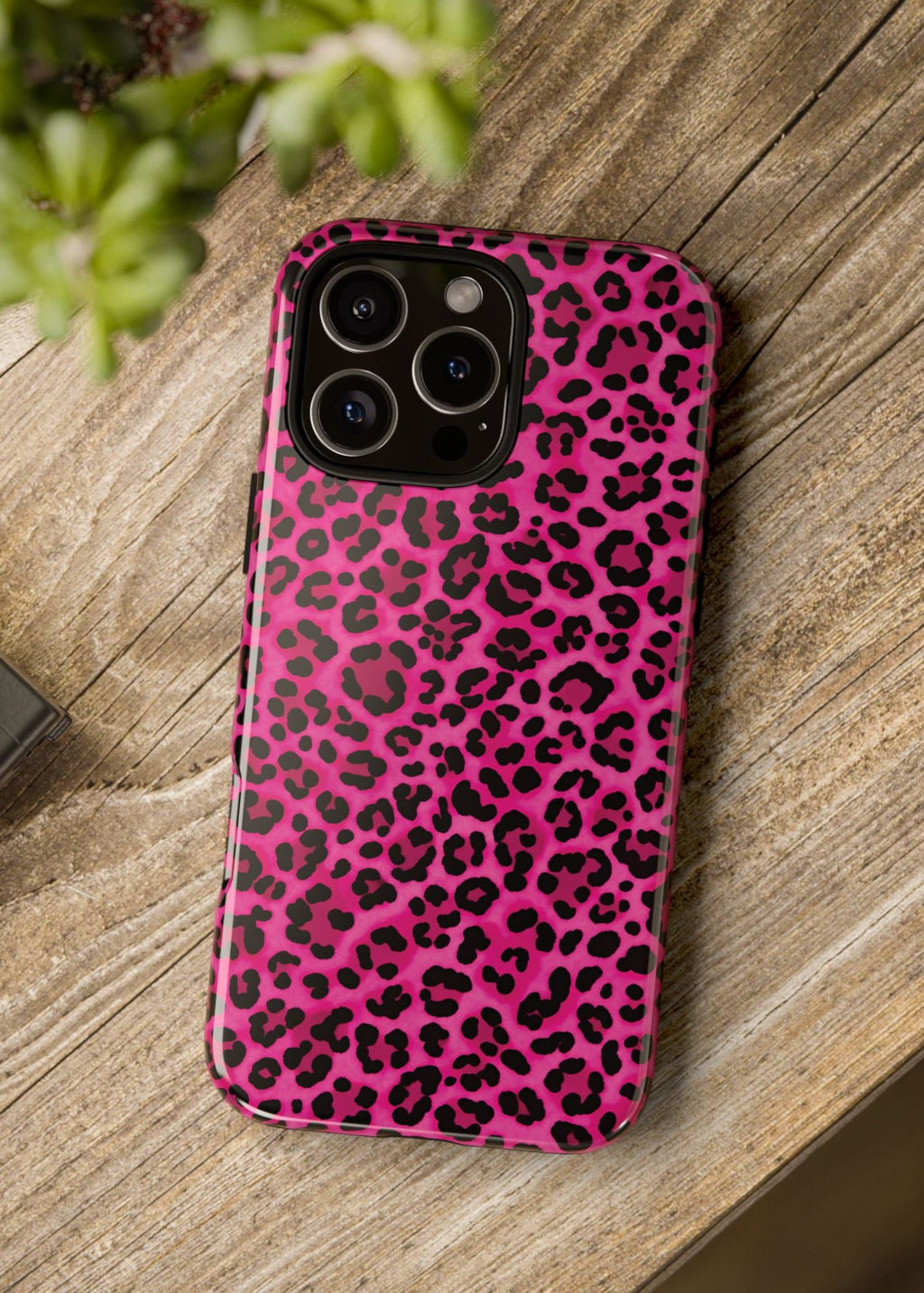 Pink leopard print phone case on a wooden surface with a camera and plant in the background